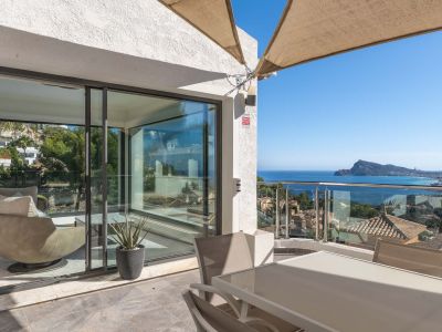 Außenterrasse mit Esstisch und Stühlen, geschützt durch ein Sonnensegel. Bietet Panoramablick auf das Meer und die Küste durch ein Glasgeländer, verbunden mit einem hellen Innenbereich.