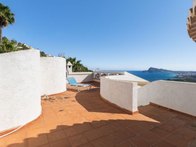 Dachterrasse mit Terrakottafliesen und weißen Strukturwänden, die einen Panoramablick auf das Meer und die Küste bietet. Eine blaue Sonnenliege lädt zum Entspannen ein.