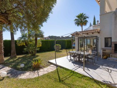 Dieser einladende Essbereich im Freien verfügt über eine geflieste Terrasse mit Metallpergola, ideal für Mahlzeiten. Ergänzt wird er durch einen gemauerten Grill und einen üppigen Garten mit Palmen und Hecke.