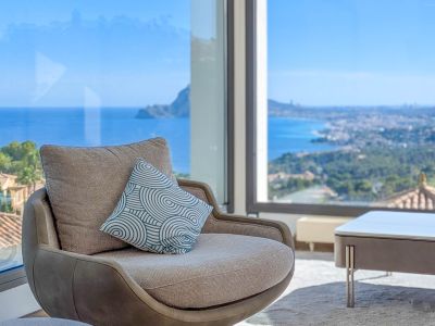 Gemütlicher Sessel am Panoramafenster mit unverbautem Blick auf das Mittelmeer und die Küstenlinie. Ein Zierkissen ergänzt das moderne Interieurdesign.