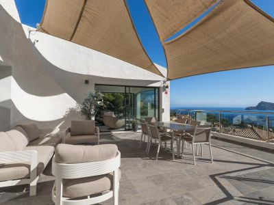 Geräumige Außenterrasse mit Lounge- und Essbereichen unter einem Sonnensegel, mit Panoramablick auf Meer und Küste durch eine Glasbrüstung.