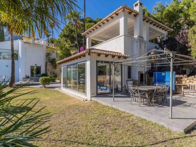 Geräumige gepflasterte Terrasse mit Essbereich unter einer Metallpergola, angrenzend an einen verglasten Raum und einen gepflegten mediterranen Garten.