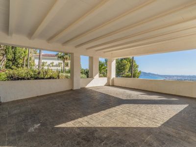 Geräumiger überdachter Carport mit gestanztem Betonboden und weißer Balkendecke. Er bietet Panoramablicke auf das Mittelmeer und die Küste, umgeben von üppiger Vegetation.