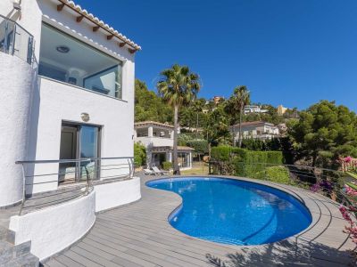 Geschwungener blauer Mosaikpool mit grauer Verbundterrasse, angrenzend an eine weiße Fassade mit Panoramafenster und Balkon mit Edelstahlgeländer.