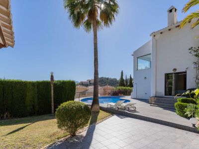 Großer Garten mit rundem Pool und Holzdeck-Solarium, begrenzt von einer weißen Villa und einer hohen Hecke. Blick auf die Hügellandschaft.
