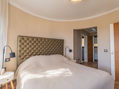 Hauptschlafzimmer mit gepolstertem Kopfteil, einer markanten geschwungenen Wand und natürlichem Licht durch transparente Vorhänge, ergänzt durch Holztüren.