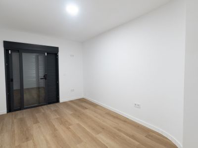 Heller Raum mit hellem Holzlaminatboden und weißen Wänden. Eine große anthrazitfarbene Glasschiebetür mit integrierten Jalousien bietet Zugang zum Außenbereich.