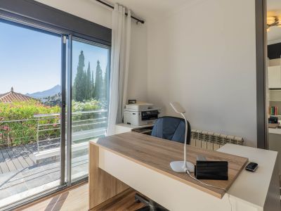 Helles Büro mit modernem Holzschreibtisch, großer Glasschiebetür zu einer Terrasse mit üppigem Grün und Panoramablick auf die Berge. Ein Spiegel reflektiert ein praktisches Bücherregal.