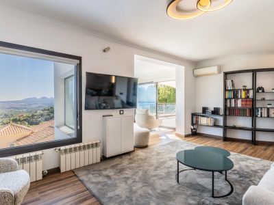 Helles Wohnzimmer mit Panoramablick auf die Berge vom Fenster und Zugang zur Terrasse mit Meerblick. Ausgestattet mit Holzboden, einem großen grauen Teppich und einem modernen Bücherregal.