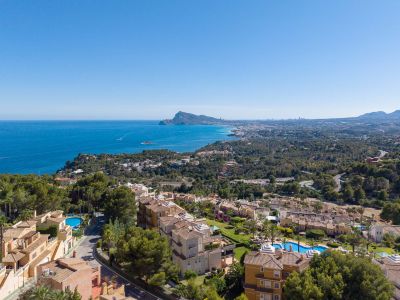 Luftaufnahme der Küste von Altea Hills, mit dem Mittelmeer, dem Peñón de Ifach und der Skyline von Benidorm in der Ferne. Terrakottadächer und Pools sind inmitten üppiger Vegetation sichtbar.