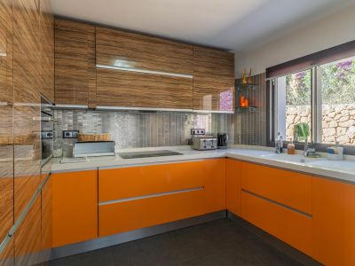 Moderne Küche mit glänzenden Holzmaser-Oberschränken und leuchtend orangefarbenen Unterschränken. Weiße Arbeitsplatte, metallische Mosaik-Rückwand und ein Fenster mit Blick auf eine Steinmauer.