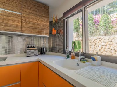 Moderne Küche mit weißer Quarz-Arbeitsplatte, leuchtend orangefarbenen Unterschränken und Oberkästen in Holzoptik. Das Fenster blickt auf eine Natursteinmauer und Pflanzen.