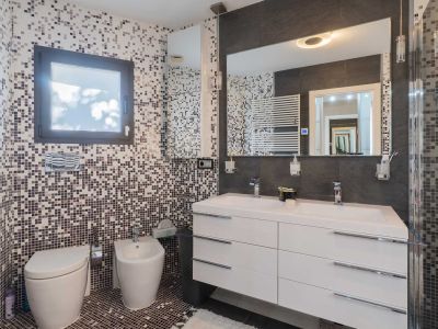 Modernes Bad mit Mosaikfliesen an Wänden und Boden in Erd-/Grautönen. Es bietet einen schwebenden Doppelwaschtisch, großen beleuchteten Spiegel, wandhängendes WC, Bidet und Handtuchwärmer.