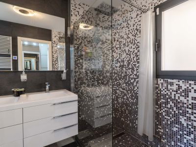 Modernes Badezimmer mit Doppelwaschtisch, weißen Unterschränken und beleuchtetem Spiegel. Die Glasdusche hat Mosaikfliesen in Grautönen und ein integriertes Fenster.