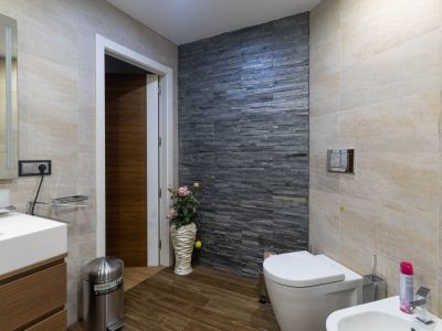 Modernes Badezimmer mit dunkler Natursteinwand, hellbeigen Fliesen und Holzoptikboden. Ausgestattet mit WC, Bidet und Waschtisch mit Waschbecken.