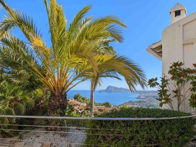 Panoramablick auf das Meer von einer Terrasse, umrahmt von üppigen Palmen. Edelstahlgeländer, niedrige Hecke und die Küste mit dem ikonischen Peñón de Ifach.