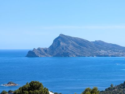 Panoramablick auf das Mittelmeer mit dem markanten Profil der Sierra Helada, einer entfernten Küstenstadt und kleinen Felseninseln unter klarem blauem Himmel.