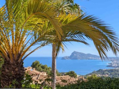 Panoramablick auf das Mittelmeer, umrahmt von Palmen, mit den Dächern von Altea Hills und dem fernen Peñón de Ifach unter blauem Himmel.