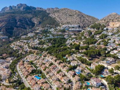 Panoramische Luftaufnahme von Altea Hills, die zahlreiche Villen mit Pools an einem grünen Hang vor einer dramatischen Bergkulisse zeigt.