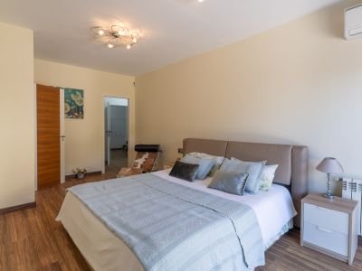 Schlafzimmer mit beigem Kopfteilbett, Laminatboden und moderner Deckenleuchte. Nachttisch mit Lampe, Heizkörper und Klimaanlage sind sichtbar.