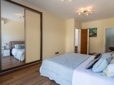 Schlafzimmer mit Bett in hellblauen Textilien, Holzlaminatboden und Schiebetürenschrank mit Spiegelfront. Eine moderne Deckenleuchte und florale Wandkunst sind sichtbar.
