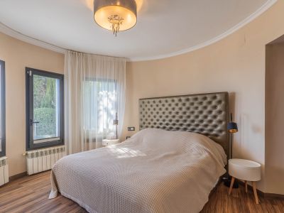 Schlafzimmer mit großem Bett und gestepptem Kopfteil. Geschwungene Wand mit dunkel gerahmten Fenstern und hellen Vorhängen. Holzboden, Heizkörper und eine moderne Deckenleuchte.