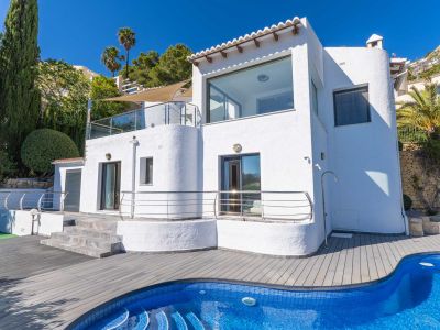 Weiße Villa mit großen Fenstern und verglasten Terrassen. Der blaue Mosaikpool und die graue Verbundterrasse werden von Zypressen und Palmen umrahmt.