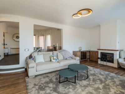 Wohnzimmer mit Kamin und rötlichem Marmorsims sowie moderner Deckenleuchte. Ein helles Sofa und tealfarbene Couchtische auf grauem Teppich führen zu einem erhöhten Bereich.