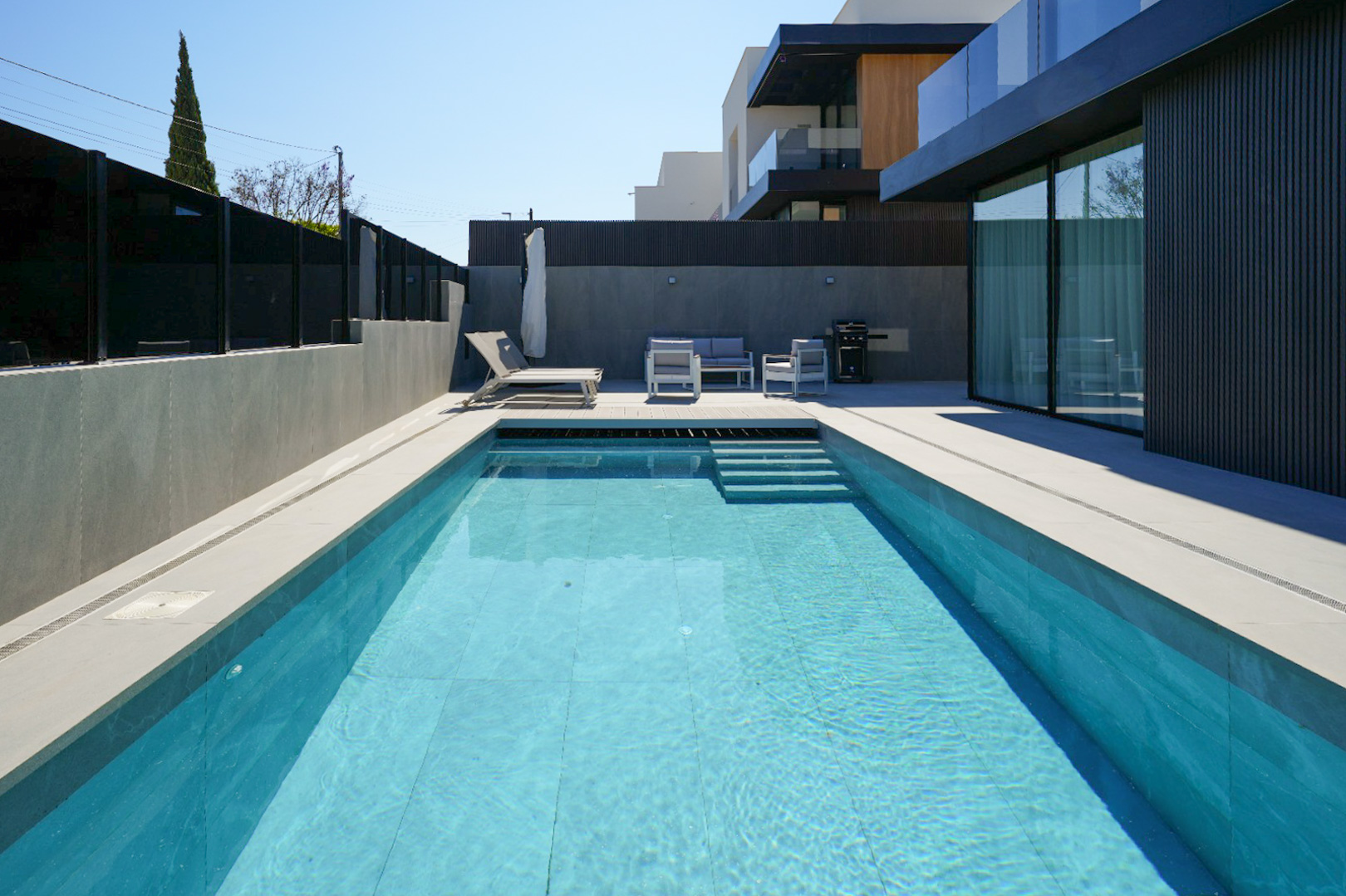 Der Außenbereich zeigt einen rechteckigen Pool mit hellblauen Fliesen und Stufen, umgeben von hellem Pflaster. Eine moderne Lounge-Gruppe und Sonnenliege laden zur Entspannung neben der eleganten Fassade ein.