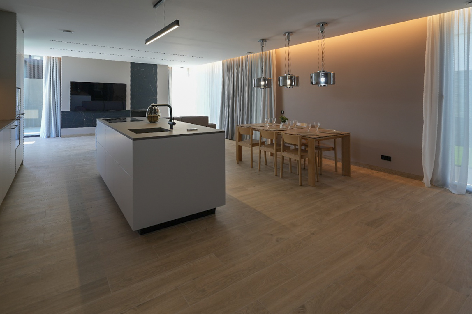 Designerküche mit zentraler Insel, integriertem Spülbecken und Kochfeld, offen zum Essbereich mit Holztisch und modernen Pendelleuchten. Heller Holzoptik-Fliesenboden.