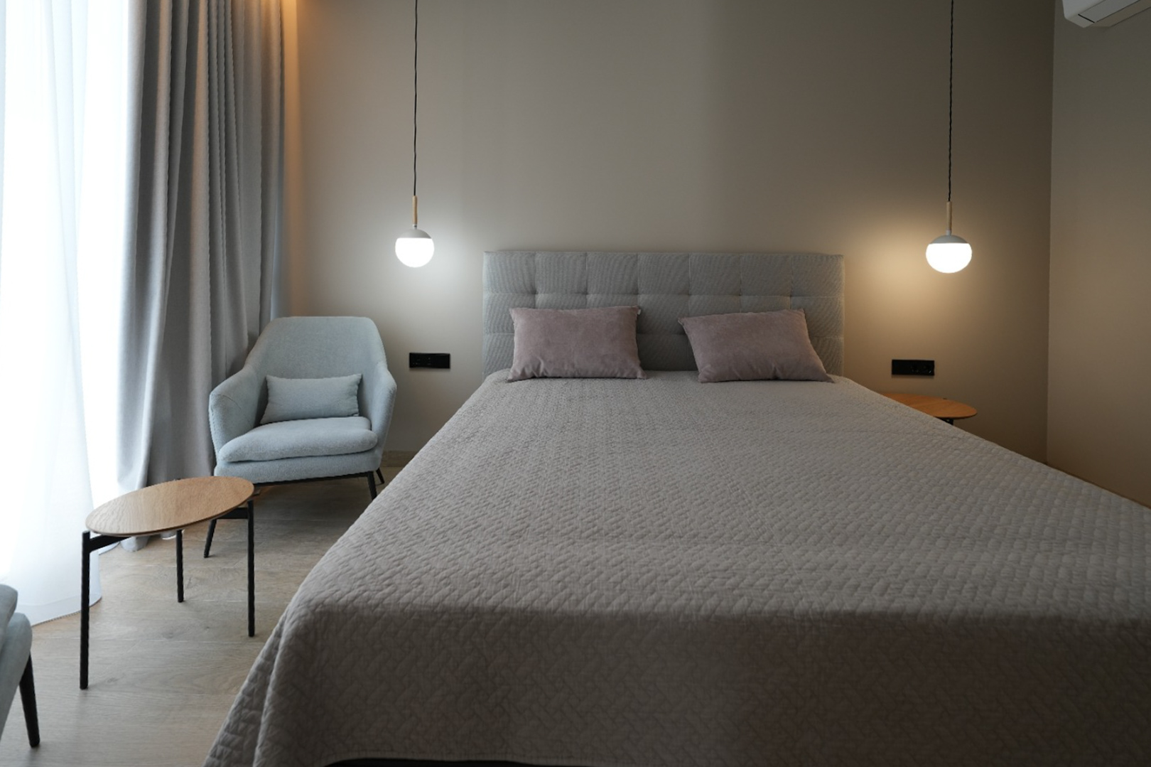 Elegantes Schlafzimmer mit grau gepolstertem Bett, zwei modernen Pendelleuchten und einem bequemen Sessel. Helle Vorhänge und Holzbodenoptik schaffen eine ruhige Atmosphäre.