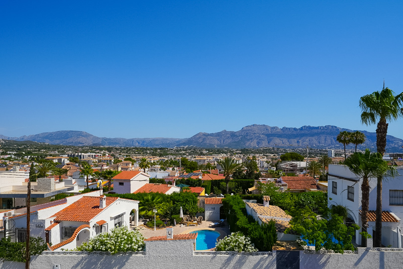 Erhöhte Ansicht des Wohngebiets von Albir, mit privatem Pool, Häusern mit Ziegeldächern und üppiger mediterraner Vegetation, vor einer Bergkulisse.