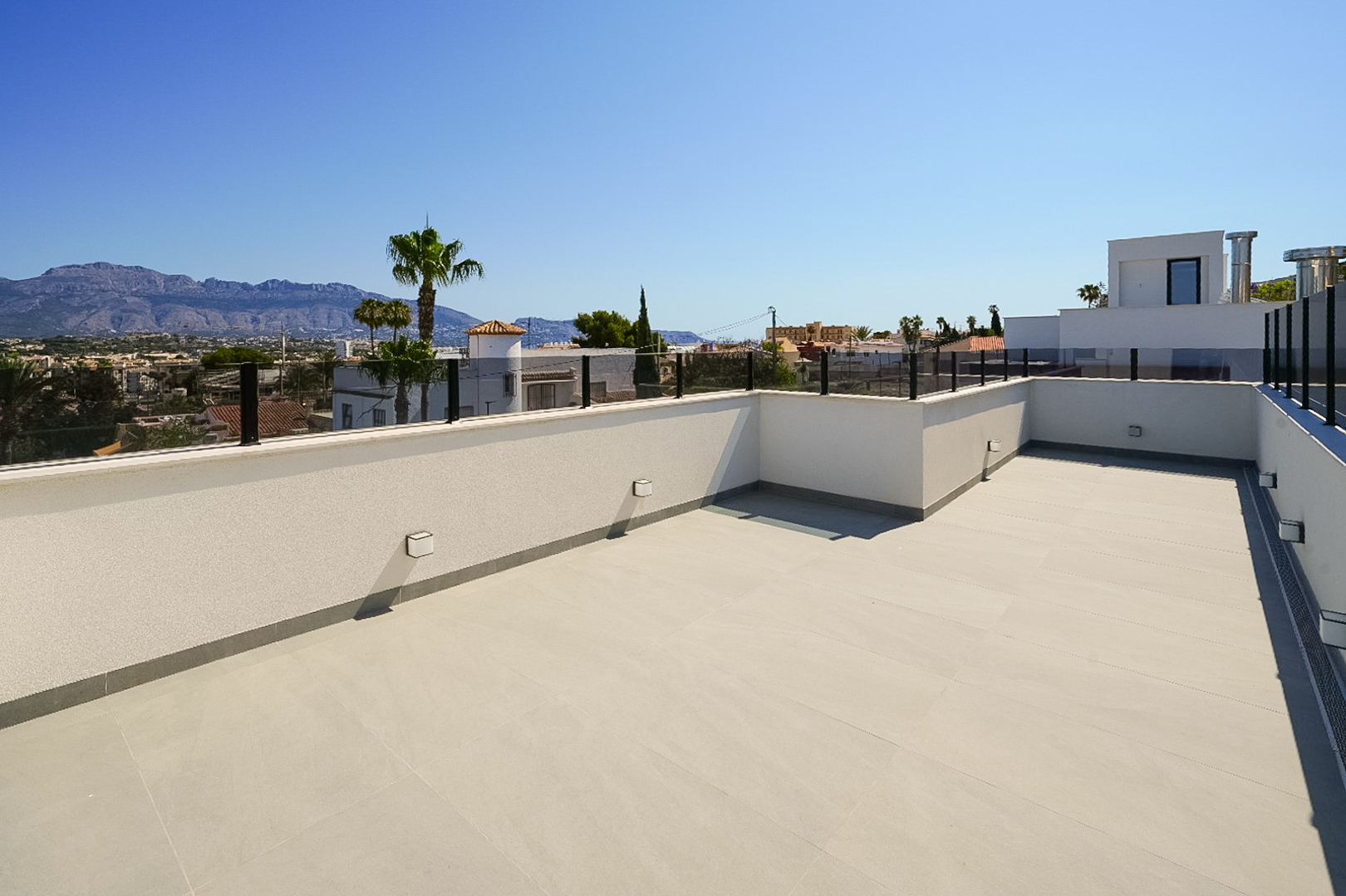 Geräumige Dachterrasse mit hellen Bodenfliesen und moderner Glasbrüstung, mit Panoramablick auf die Berge und die umliegende Stadt.