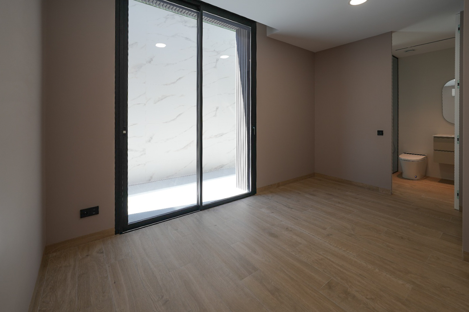 Geräumiges Zimmer mit Holzoptikfliesen und hellen Wänden. Große schwarze Glasschiebetüren führen zu einem Außenbereich mit weißen Marmorfliesen. Integriertes Badezimmer sichtbar.