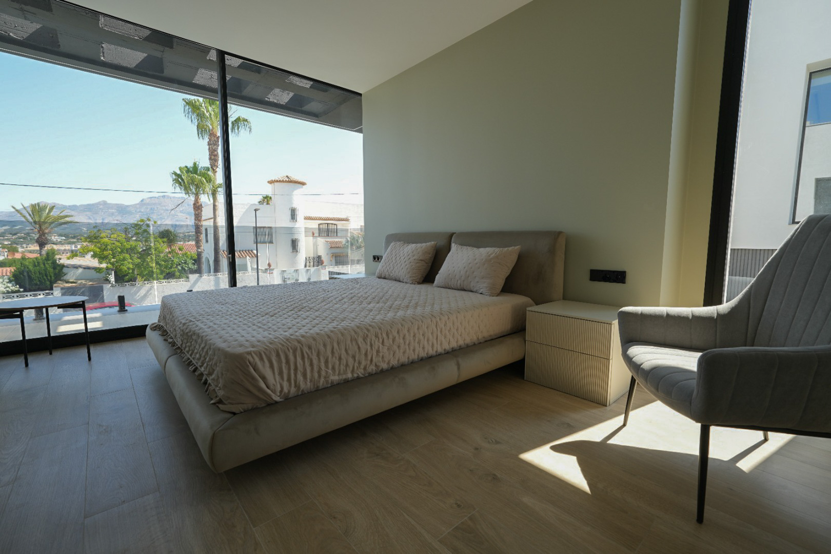 Hauptschlafzimmer mit gepolstertem Bett, geripptem Nachttisch und raumhohen Fenstern mit Blick auf Palmen und Berge.