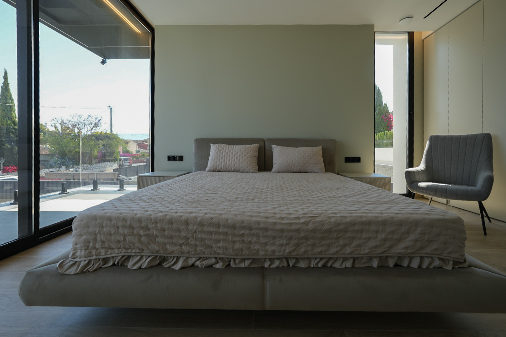 Hauptschlafzimmer mit schwebendem Bett, gepolstertem Kopfteil, großen Fenstern mit Meerblick und Sessel. Integrierte Nachttische.