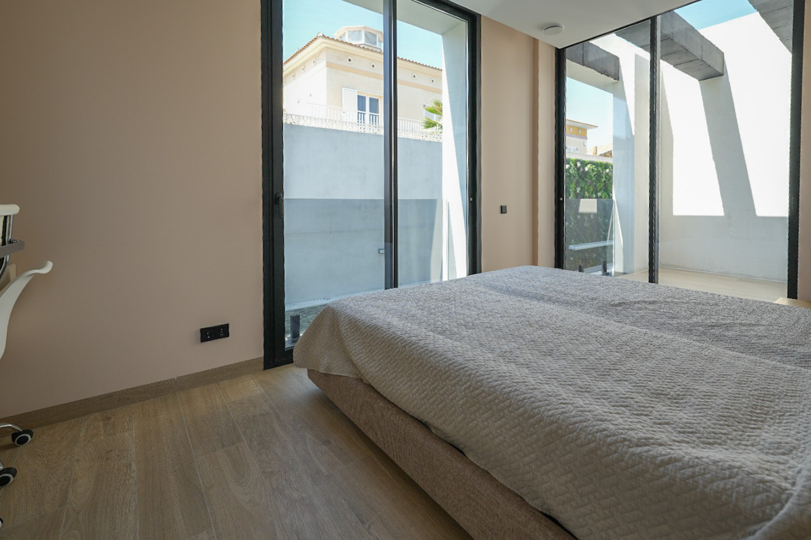 Helles Schlafzimmer mit hellem Holzfußboden und großen, schwarz gerahmten Glasschiebetüren, die den Blick auf ein Nachbarhaus und eine grüne Wand freigeben.