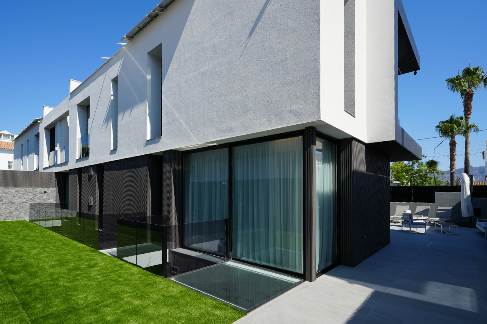 Moderne Außenansicht der Villa mit kontrastierender weiß-grauer und dunkler Fassade, großen Glasfronten, Kunstrasenfläche und gefliester Terrasse mit Sitzgelegenheiten.