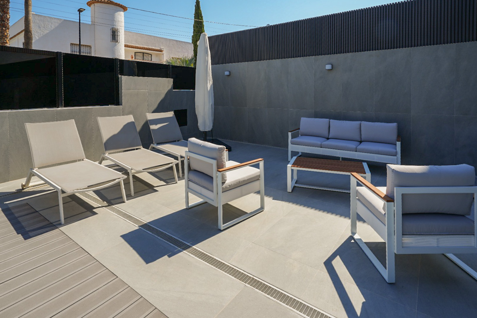 Moderne Außenterrasse mit Lounge-Möbeln in Weiß und Grau, kombiniertem Feinsteinzeug- und WPC-Boden. Sonnenliegen und eine Sitzgruppe sind vorhanden.