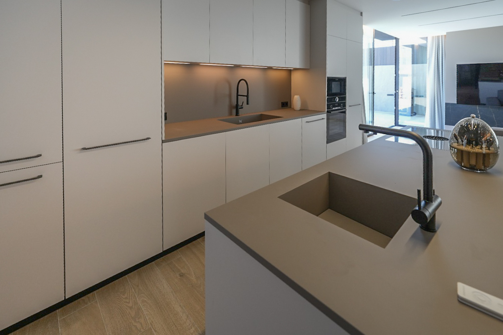 Moderne Küche mit Kücheninsel und integriertem Doppelwaschbecken. Minimalistische Schränke in neutralen Tönen, Einbaugeräte und heller Holzfußboden, mit Zugang zum Außenbereich.
