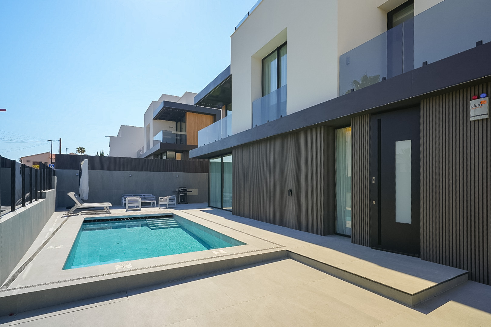Moderner Villa-Außenbereich mit rechteckigem Pool und integrierten Stufen, umgeben von hellem Pflaster. Dunkle Lamellenverkleidung der Erdgeschossfassade und markante Eingangstür.