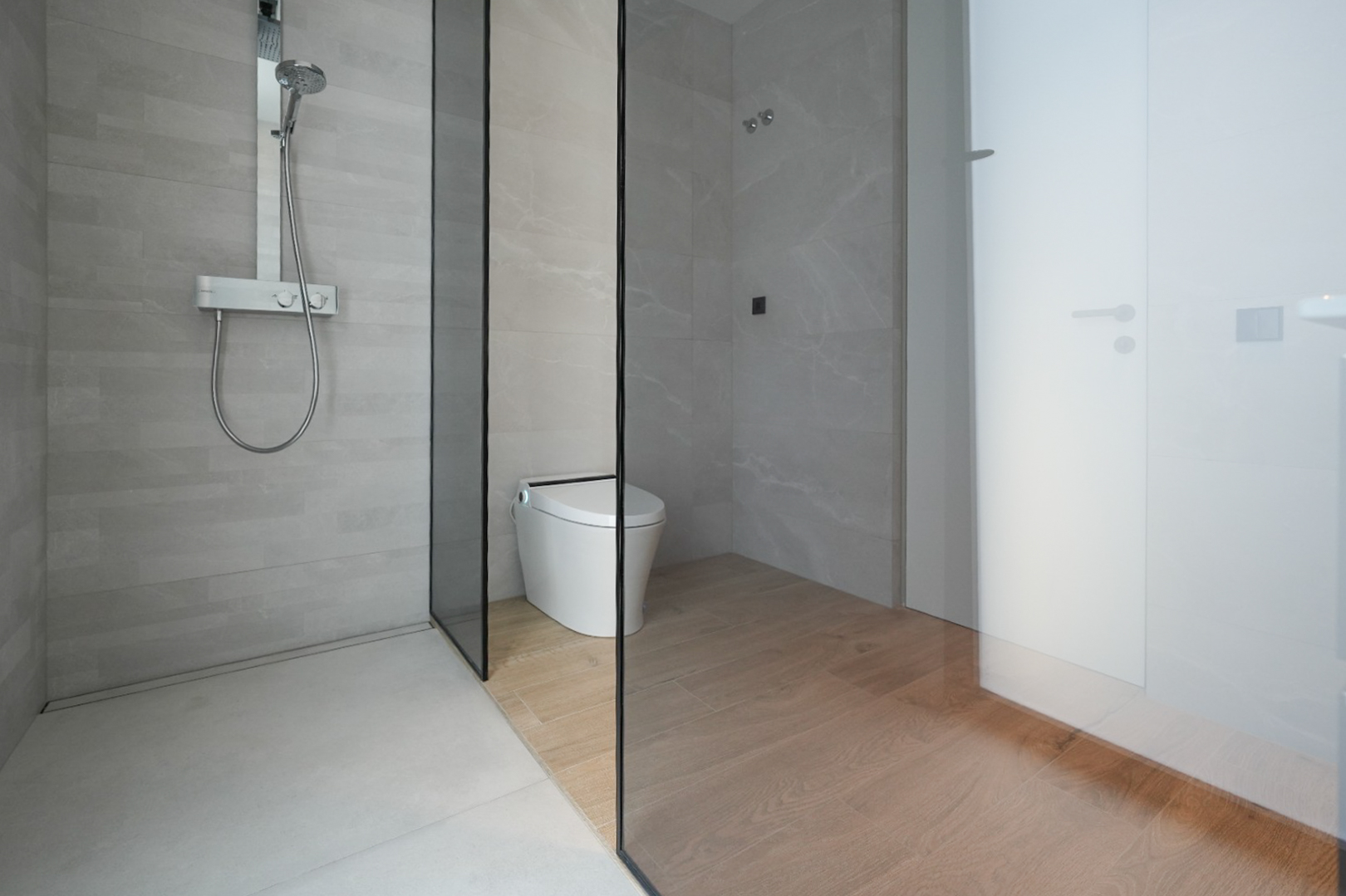 Modernes Badezimmer mit ebenerdiger Dusche, hellgrauen Fliesen und verchromter Thermostatarmatur. Eine Glaswand trennt die Dusche vom Smart-WC-Bereich mit Holzoptik-Fliesenboden.