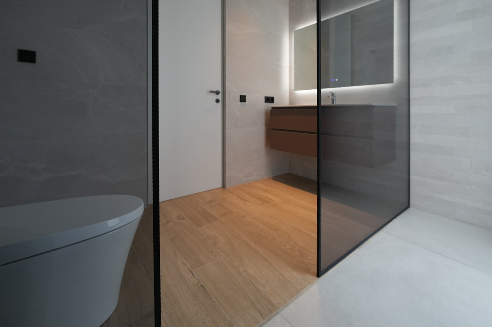 Modernes Badezimmer mit wandhängendem WC, schwebendem Holzwaschtisch, beleuchtetem Spiegel und Glasduschabtrennung. Holzbodenfliesen ergänzen die grauen Wand- und Duschfliesen.