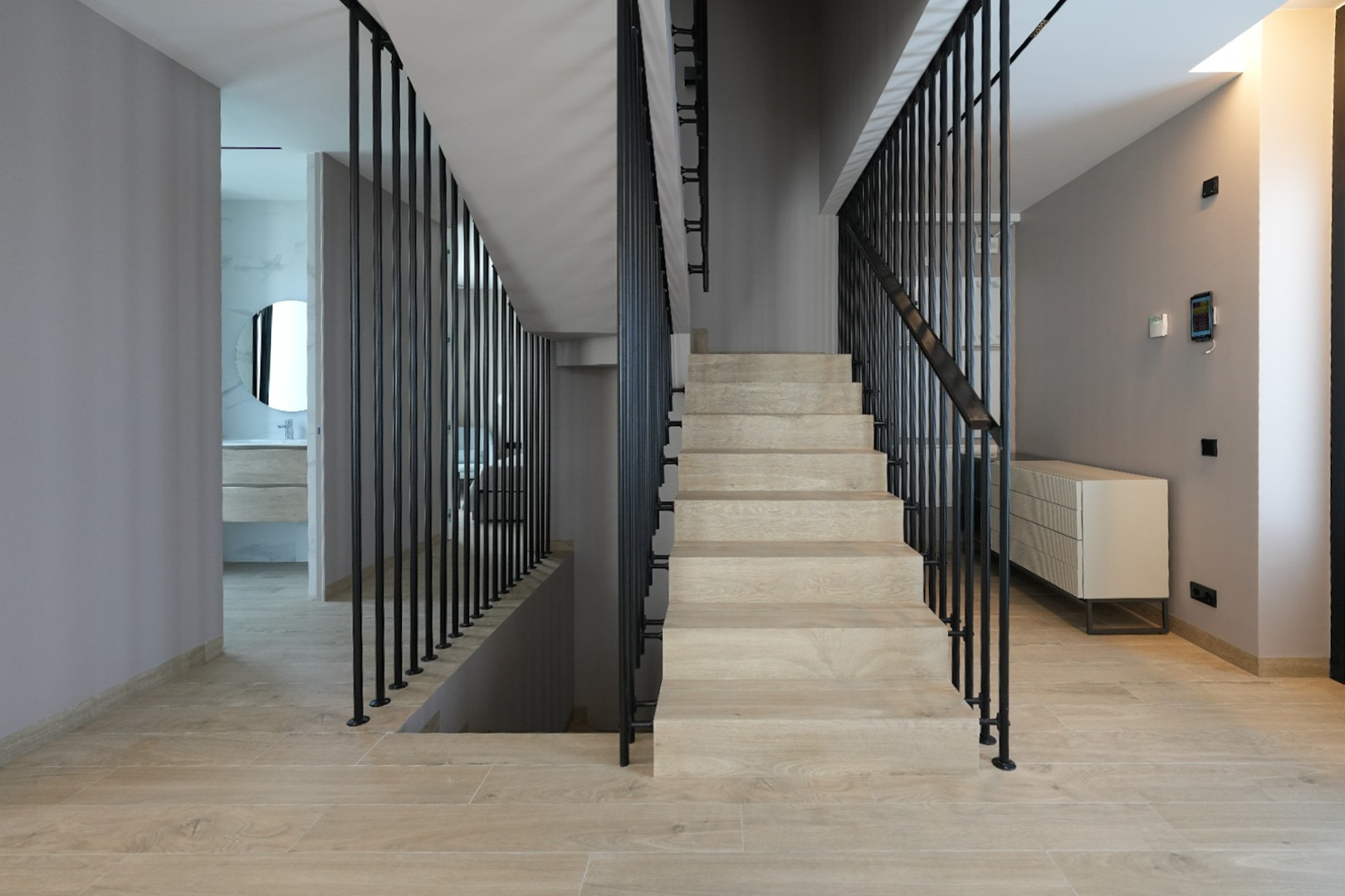 Modernes Interieur mit heller Holztreppe und schlankem schwarzen Vertikalgeländer. Ein Smart-Home-Bedienfeld ist sichtbar, führt zu einem Bad mit Rundspiegel.