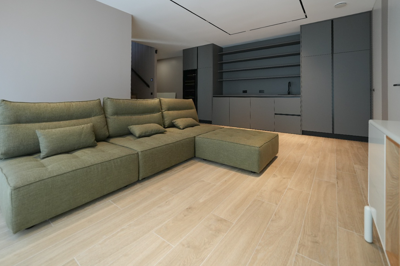 Olivgrünes Modulsofa in offenem Wohnbereich mit hellem Holzoptik-Fliesenboden. Im Hintergrund dunkle Einbauschränke mit Spüle und Regalen.