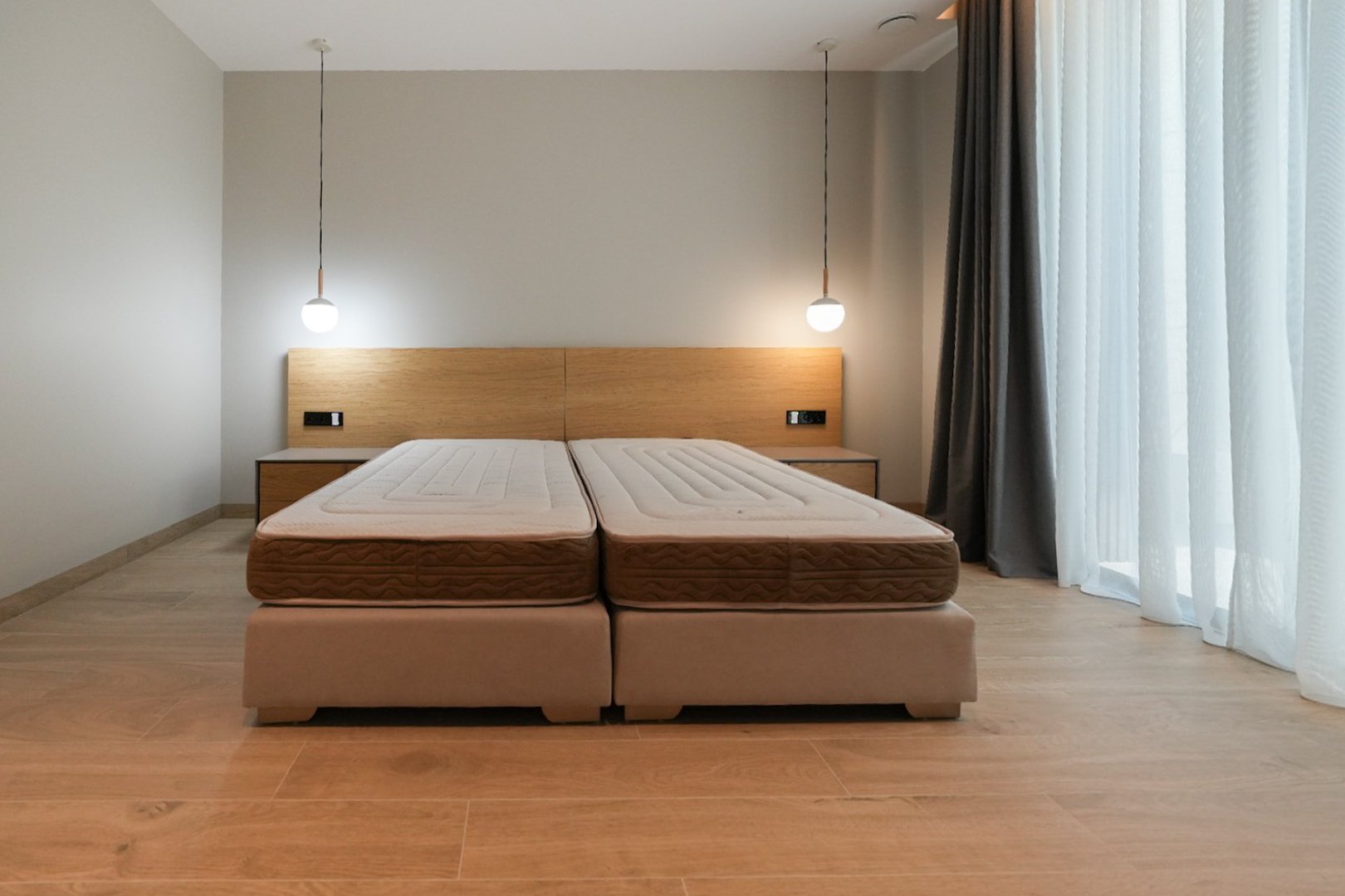 Schlafzimmer mit Doppelbett aus zwei Polsterbetten, hellem Holzkopfteil mit integrierten Nachttischen und zwei modernen Pendelleuchten. Großes Fenster mit Vorhängen.