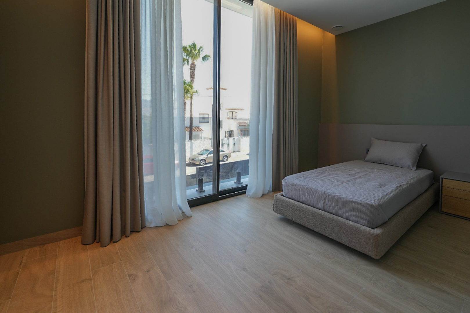Schlafzimmer mit Einzelbett, hellem Holzfußboden und olivgrünen Wänden. Große Fenster mit Doppelvorhängen bieten Blick auf die Straße und Palmen.