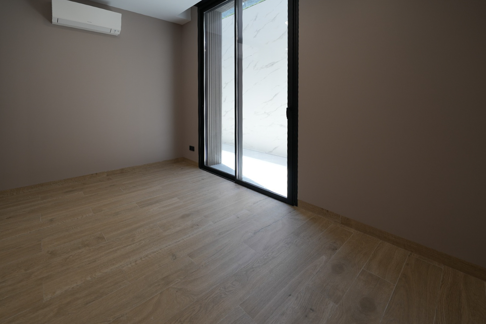 Unmöblierter Raum mit hellem Holzfliesenboden und taupefarbenen Wänden. Eine schwarze Glasschiebetür und eine Wandklimaanlage sind sichtbar.