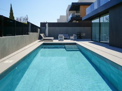 Der Außenbereich zeigt einen rechteckigen Pool mit hellblauen Fliesen und Stufen, umgeben von hellem Pflaster. Eine moderne Lounge-Gruppe und Sonnenliege laden zur Entspannung neben der eleganten Fassade ein.