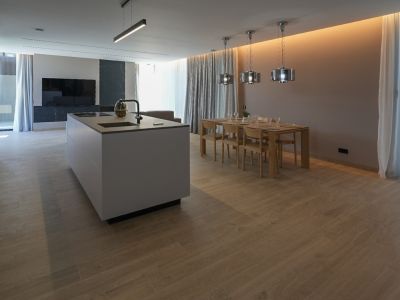 Designerküche mit zentraler Insel, integriertem Spülbecken und Kochfeld, offen zum Essbereich mit Holztisch und modernen Pendelleuchten. Heller Holzoptik-Fliesenboden.